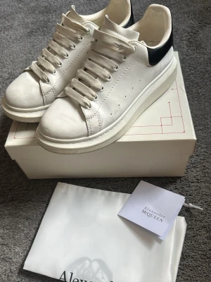 Alexander McQueen vita sneakers - Stilrena vita sneakers från Alexander McQueen med svart hälparti och guldfärgad logga. Skorna har tjock sula, perforerade detaljer på sidorna och är tillverkade i läder. Klassisk låg modell med vita skosnören och exklusiv känsla. Dustbag o box medföljer! Skorna är lite smutsiga på undersidan!