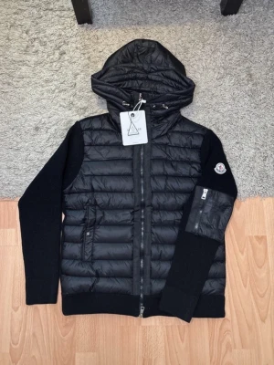 Svart cardigan med huva från Moncler - Snygg svart cardigan från Moncler med stickade ärmar och vadderad kropp. Jackan har huva, dragkedja framtill och en praktisk ficka med dragkedja på ärmen. Moncler-logga på vänster ärm. Perfekt för kalla vinterdagar.