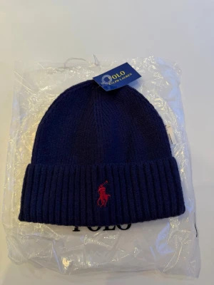 Mörkblå mössa/beanie från Polo Ralph Lauren - Mörkblå ribbstickad mössa från Polo Ralph Lauren med uppvikt kant och röd broderad logga framtill. Klassisk och stilren design som passar perfekt till kalla dagar. Tillverkad i mjukt material för extra komfort.  samma dag                                                                          📦Fraktas samma dag📦 ⭐️Helt ny samt oanvänd⭐️ Har du frågor eller funderingar så hör gärna av dig🤗