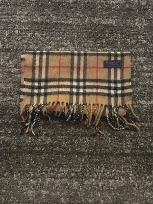 Burberrys rutig halsduk i lammull - Klassisk halsduk från Burberrys i beige, svart, vitt och rött rutmönster. Tillverkad i 100% lammull och har fransar i ändarna. Perfekt accessoar för att lyfta din outfit med en tidlös brittisk stil.