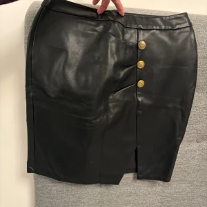 Svart skinnkjol med guldknappar - Snygg svart kjol i skinnimitation med tre dekorativa guldknappar framtill och en slits. Kjolen har en rak passform och är perfekt för att lyfta vilken outfit som helst.Chiara Forthi.använd 1 gång.Har flera kjolar till salu skicka gärna pm. Säljer väldigt mycket tjej damkläder klänningar gör bra paketpris 