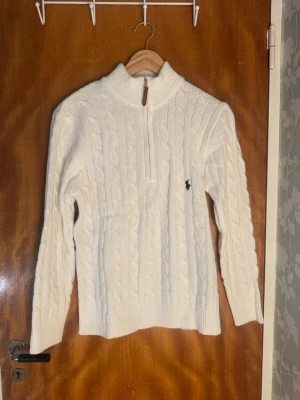 Vit kabelstickad tröja Polo Ralph Lauren - Vit kabelstickad tröja från Polo Ralph Lauren med halv dragkedja och hög krage. Tröjan har lång ärm, ribbstickade muddar och den klassiska broderade loggan på bröstet. Perfekt för en stilren och klassisk look.