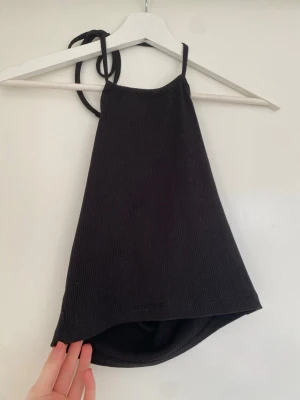 Svart ribbad linnetopp med knyt i nacken. - Svart ribbad linnetopp med knyt i nacken. Toppen är ärmlös och har en croppad passform som ger en snygg siluett. Perfekt för att styla med jeans eller kjol. Har en öppen rygg perfekt för sommaren eller fest.