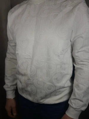 Vit stickad tröja från Hugo Boss - Vit stickad tröja från Hugo Boss med diskret mönster av stora bokstäver 'HUGO' över hela plagget. Tröjan har rund halsringning och ribbade muddar vid ärmslut och nederkant. Perfekt för en stilren och modern look.