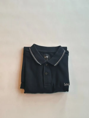 Mörkblå pikétröja från Lee - Klassisk mörkblå pikétröja från Lee med vit piping på kragen och ärmsluten. Tröjan har korta ärmar, knappslå med två knappar och diskret Lee-logga broderad på bröstet. Perfekt till jeans eller shorts för en clean look.