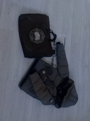 Mörkgrå dunjacka med huva från Canada Goose - Snygg mörkgrå dunjacka från Canada Goose med stor huva och diskret logga på ärmen. Jackan har quiltad design, dragkedja och knappar framtill samt två stora fickor och en inuti. Perfekt för kalla dagar.