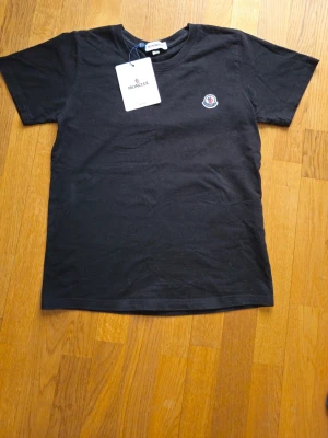 Svart Moncler t-shirt i bomull - Svart t-shirt från Moncler med klassisk rund hals och liten Moncler-logga på bröstet. Tillverkad i mjuk bomull och har en enkel, stilren design som passar till det mesta. Perfekt för dig som gillar exklusiva och snygga basplagg.