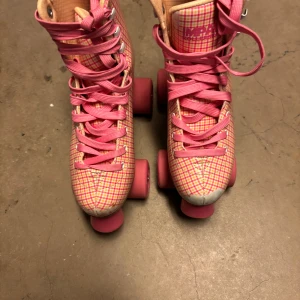 Rosa rutiga rullskridskor - Säljer ett par retroinspirerade rullskridskor i rosa med rutigt mönster och rosa hjul. Skorna har klassisk snörning och detaljer i beige samt motiv på sidan. Perfekta för dig som vill sticka ut på rullbanan! Pris kan diskuteras vid snabb affär