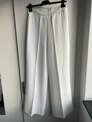 Vita vida kostymbyxor med pressveck - Snygga vita kostymbyxor med hög midja och vida ben. Byxorna har klassiska pressveck framtill och en stilren design utan synliga fickor. Perfekta för en clean och modern look. Materialet ser ut att vara ett mjukt tyg med lite fall.