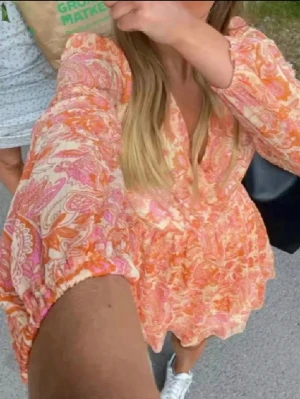 Blommig omlottklänning i orange och rosa - Säljer en kort omlottklänning med blommigt mönster i orange, rosa och vitt. Klänningen har v-ringning, volangdetaljer och långa ärmar med lätt puff. Perfekt för sommarens alla tillfällen!