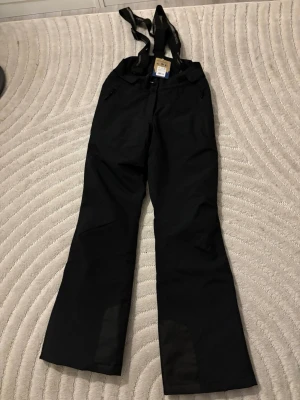 Svarta täckbyxor Stormberg XS - Svarta täckbyxor från Stormberg med justerbara hängslen och dragkedjor vid fickorna. Tillverkade i slitstarkt, vattenavvisande material och fodrade för extra värme.