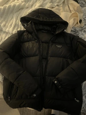 Svart pufferjacka från Alessandro Zavetti - Säljer en svart pufferjacka från Alessandro Zavetti med huva och dragkedja framtill. Jackan har två fickor med dragkedja och en snygg logga på bröstet. Oversized look och riktigt skön för kalla dagar. Perfekt för dig som vill ha en stilren och varm vinterjacka.