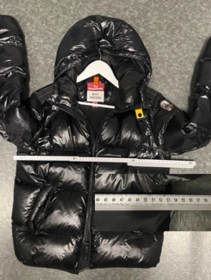 Svart pufferjacka från Parajumpers - Snygg svart pufferjacka från Parajumpers med glansig finish, huva och två stora fickor framtill. Jackan har märkets klassiska patch på ärmen och gul detalj vid halsen. Perfekt för kalla vinterdagar och riktigt varm tack vare det tjocka materialet.