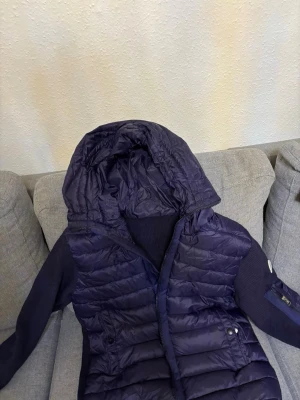 Mörkblå dunväst från Moncler - Snygg mörkblå dunväst från Moncler med huva och stickade detaljer på sidorna. Västen har dragkedja framtill, två fickor med tryckknappar och Moncler-logga på ärmen. Perfekt för lager-på-lager och streetstyle.