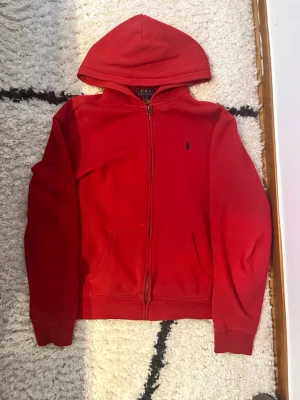 Röd hoodie från Polo Ralph Lauren - Riktigt snygg röd hoodie från Polo Ralph Lauren med dragkedja framtill och klassisk huva. Har två fickor på magen och den ikoniska lilla loggan broderad på bröstet. Perfekt för en chill och stilren look. Mjuk bomullskänsla och ribbade muddar vid ärmar och midja. Står XL men det passar S och XS