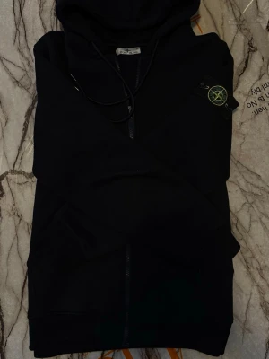 Svart hoodie från Stone Island - Svart hoodie från Stone Island med dragkedja framtill och klassisk logotyp-patch på vänster ärm. Tröjan har huva med dragsko och två fickor framtill. Perfekt för en stilren och avslappnad look.