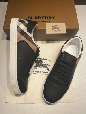 Burberry svarta sneakers med rutigt mönster - Snygga svarta sneakers från Burberry med klassiskt beige, svart och vitt rutmönster på hälen. Skorna har vita sulor, svarta skosnören och detaljer i läder och textil. Perfekta för dig som vill ha en stilren och lyxig look med ikoniska Burberry-detaljer.