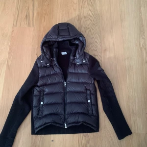 Svart cardigan med huva från Moncler - Snygg svart cardigan från Moncler med stickade ärmar och huva. Västen har dragkedja framtill, två fickor med tryckknappar och Moncler-logga på ärmen. Materialmix av dun och stickat ger en modern look.