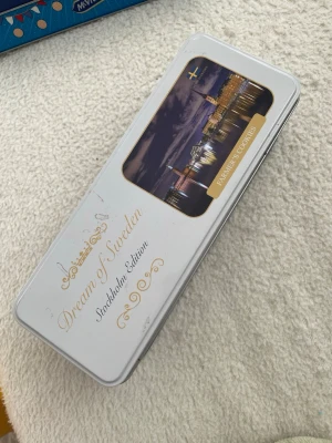 Plåtburk Dream of Sweden Stockholm - Säljer en rektangulär plåtburk från Dream of Sweden, Stockholm Edition. Burken är vit och blå med gulddetaljer och har ett foto av Stockholms stad på locket. Texten 'Farmer Cookies' finns på framsidan. Perfekt som souvenir eller förvaring.