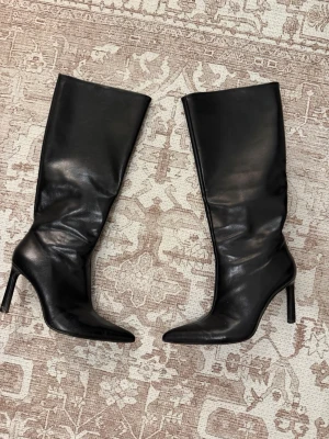 Svarta höga boots med klack - Snygga svarta stövlar i skinnimitation med spetsig tå och hög smal klack. Stövlarna går upp till knäet och har dragkedja på insidan för enkel påtagning. Perfekta för att lyfta vilken outfit som helst.