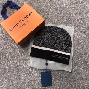 Svart Louis med vit text - Snygg svart mössa med vit logga och klassiskt monogrammönster i grått och vitt. Mössan har en bred svart kant med vit rand och text broderad framtill. Perfekt accessoar för dig som gillar exklusiv streetstyle.