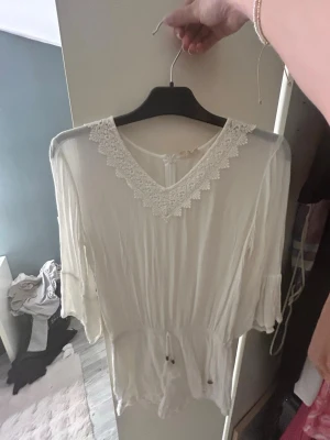 Vit blus med spetsdetalj från Ü - Söt vit jumpsuit från Ü med v-ringning och spetsdetaljer runt halsen. Blusen har trekvartsärm och dragsko i midjan för en snygg passform. Tunt och luftigt material som känns skönt mot huden.