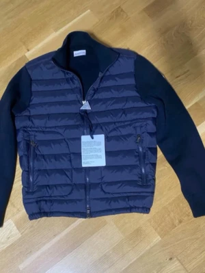 Mörkblå dunväst med ärmar - Snygg mörkblå dunväst med quiltad framsida och stickade ärmar. Västen har dragkedja framtill, två fickor med dragkedja och ribbad nederkant. Perfekt för lager-på-lager och en sportig look. Pris kan diskuteras, äkta jacka storleken är S