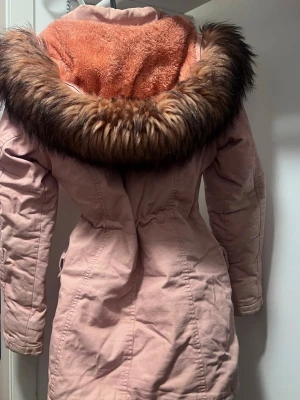 Ljusrosa parkas med fuskpäls - Snygg ljusrosa parkas med stor huva fodrad i mjuk orange teddy och kantad med brun fuskpäls. Jackan har dragsko i midjan, flera fickor med knappar och dragkedja framtill. Perfekt för kalla dagar och ger en cool streetstyle-vibe.
