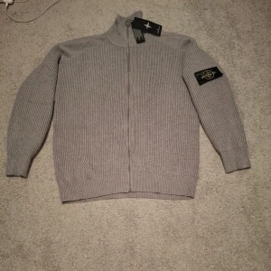 Grå ziptröja från Stone Island - Säljer en grå ribbstickad ziptröja från Stone Island med hög krage och ikonisk patch på ärmen. Tröjan har hel dragkedja framtill och ribbade muddar vid ärmslut och nederkant. Perfekt för dig som gillar stilrena och exklusiva plagg.