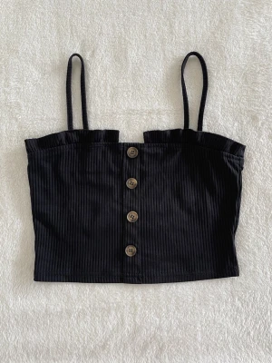 Svart ribbad crop top med knappar - Svart croppad linnetopp med smala axelband och ribbad struktur. Toppen har dekorativa knappar framtill och en lätt rynkad kant upptill för en trendig look. I fint skick, ej genomskinlig