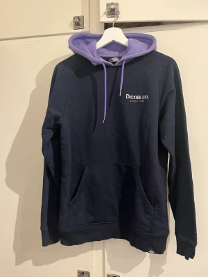 Mörkblå hoodie från Dickies - Snygg mörkblå hoodie från Dickies med lila huva och matchande dragsko. Framtill finns en vit Dickies Co.-logga och en stor känguruficka. Hoodien har ribbade muddar vid ärmar och nederkant. Perfekt för en chill och avslappnad stil. Djur och rökfritt hem.