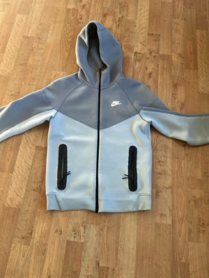 Grå Nike hoodie med dragkedja - Snygg grå hoodie från Nike med dragkedja framtill och två svarta dragkedjeförsedda fickor. Hoodien har en klassisk huva och Nike-logga på bröstet. Tillverkad i mjukt material som passar perfekt till en avslappnad streetwear-look.