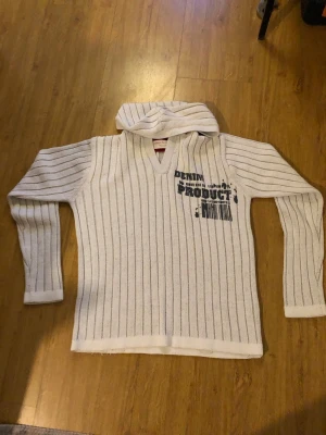 Vintage hoodie Jack and Jones  - Vit hoodie med tunna svarta vertikala ränder och tryck på bröstet från Denim Product. Tröjan har huva, lång ärm och en avslappnad passform. Materialet ser ut att vara stickat och mjukt, perfekt för en chill och stilren look.