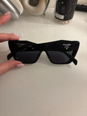 Svarta cat eye solglasögon från Prada - Snygga svarta solglasögon från Prada i cat eye-modell med breda skalmar och mörka glas. Modern look med tydlig Prada-logga på båda sidor. Perfekta för dig som vill sticka ut med en lyxig accessoar. (Obs köpa i andra hand så kan inte garantera att de är äkta, finns heller ingen box till)