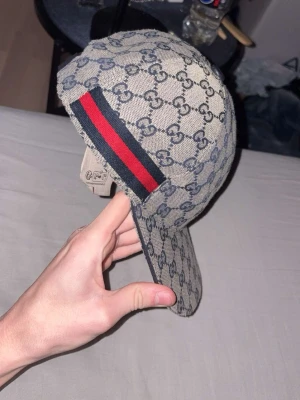Gucci keps med GG-mönster - Säljer en snygg Gucci keps i beige med klassiskt GG-monogram över hela kepsen. På sidan finns en ikonisk röd och marinblå rand. Kepsen har böjd skärm och justerbar rem bak. Materialet är canvas med mörkblå insida. Perfekt för dig som gillar lyxiga accessoarer.
