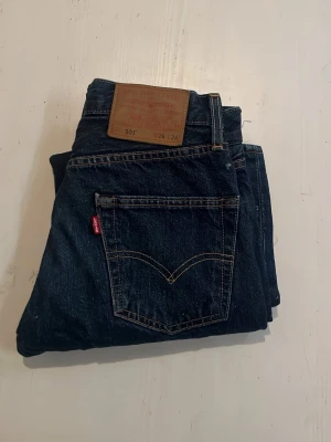 Levi's 501 mörkblå jeans W26 L28 - Klassiska Levi's 501 jeans i mörkblå denim med raka ben och femficksmodell. Snygga kontrastsömmar och ikonisk röd Levi's-tag på bakfickan. Tillverkade i slitstarkt bomullsmaterial och har läderpatch bak i midjan.