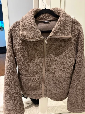 Beige teddyjacka med dragkedja - Mysig beige teddyjacka från Lager 157  med hög krage och dragkedja framtill. Jackan har två stora fickor fram och är gjord i fluffigt, mjukt teddy-material som håller dig varm. Perfekt för kyliga dagar och ger en avslappnad, trendig look.