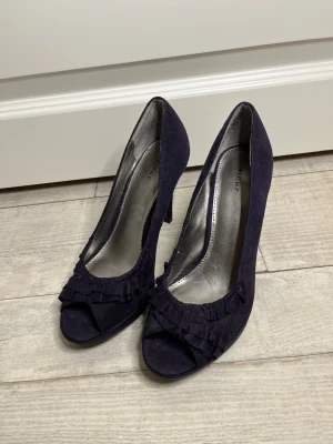 Mörklila mocka-pumps med volang - Snygga mörklila pumps från Fioni i storlek 38 med mocka och öppen tå och volangdetaljer framtill. Klackarna är smala och eleganta, perfekta för att lyfta din outfit. Insidan är fodrad i silverfärgad syntet för en cool kontrast.