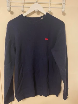 Mörkblå stickad Levi's tröja - Mörkblå stickad tröja från Levi's med rund halsringning och diskret röd Levi's-logga på bröstet. Tröjan har långa ärmar och ribbade muddar vid ärmslut och nederkant. Perfekt för kalla dagar och enkel att matcha med olika outfits.