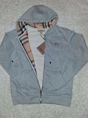 Grå hoodie från Burberry med dragkedja - Snygg grå hoodie från Burberry med klassiskt rutigt foder i huvan och dragkedja framtill. Broderad logga på bröstet och två fickor. Perfekt för en stilren och avslappnad look. Tillverkad i mjukt bomullsmaterial.