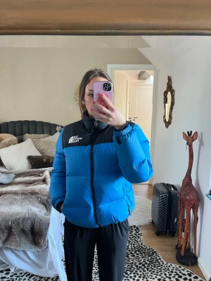 Blå pufferjacka från The North Face - Säljer en blå pufferjacka från The North Face med svarta detaljer vid axlar och krage. Jackan har en hög krage, dragkedja framtill och logga på bröstet. Perfekt för kalla vinterdagar eller skidåkning. Storlek XL youth/junior men sitter som en dam S/M