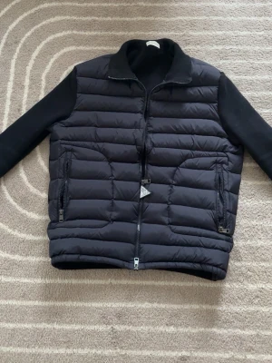 Svart dunväst från Moncler - Snygg svart dunväst från Moncler med quiltad framsida, dragkedja och två fickor med dragkedja. Ärmar och krage i stickat material ger en cool kontrast till den glansiga dunytan. Perfekt för lager-på-lager och streetstyle. Den är storlek XL men sitter som L och M på Moncler så är storlekerna skillnad.