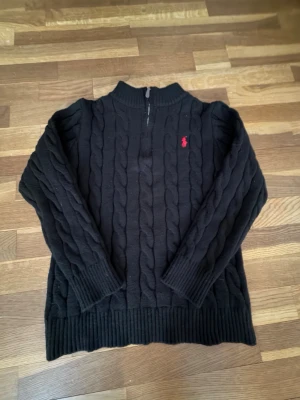 Svart kabelstickad tröja Polo Ralph Lauren - Kabelstickad svart tröja från Polo Ralph Lauren med halv dragkedja och röd broderad logga på bröstet. Tröjan har ribbade muddar vid ärmslut och nederkant samt hög krage. Perfekt för en stilren och klassisk look.