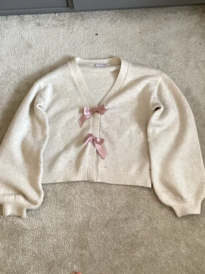 Beige kofta med rosa rosetter - Supersöt beige kofta med v-ringning och två rosa sidenrosetter framtill. Koftan har ballongärmar. 💘💘💝💝