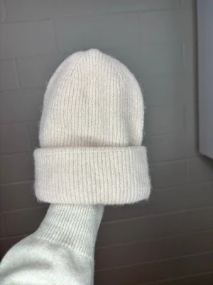 Vit ribbstickad mössa - Säljer en stilren vit ribbstickad mössa med uppvikt kant. Mössan är mjuk och har en klassisk form som passar perfekt till kalla dagar. Materialet ser ut att vara ull eller en ullmix för extra värme och komfort.