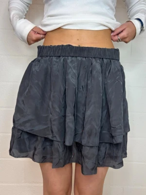 Mörkgrå volangkjol med resårmidja - Säljer en mörkgrå kortkjol med dubbla volanglager och resår i midjan. Kjolen har en luftig och flowy känsla, perfekt för att styla med en enkel topp eller stickad tröja. Materialet känns lätt och mjukt mot huden.