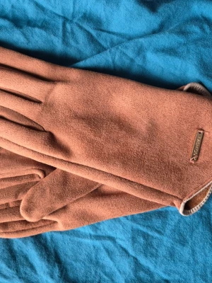 Bruna mockahandskar  - Snygga bruna handskar i mjuk mocka. De har en klassisk och stilren design med slät yta och diskret detalj vid handleden. Perfekta för att hålla händerna varma under kyliga dagar.