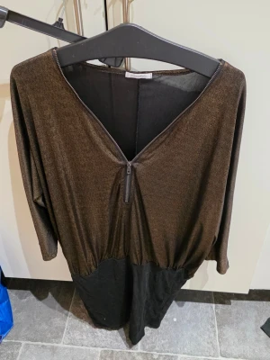 Svart och brun långklänning med zip - Cool långklänning i svart och brun med glittrig finish. Klänningen har v-ringning, lång ärm och en dekorativ dragkedja framtill. Materialet känns mjukt och stretchigt, perfekt för dig som vill sticka ut lite extra.