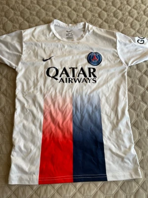 PSG matchtröja Nike Dri-FIT - Vit PSG matchtröja från Nike med Dri-FIT-material. Framsidan har röda och blå vertikala ränder, PSG-logga på bröstet och Qatar Airways-tryck. Perfekt för fotbollsträning eller att bära som supporter.