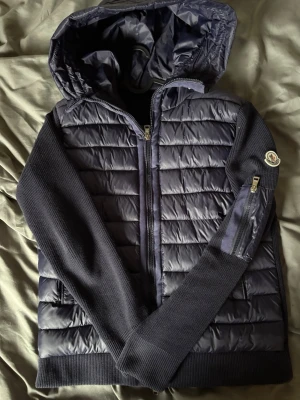 Moncler cardigan Mörkblå  - Snygg mörkblå dunjacka från Moncler med quiltad front och rygg, stickade ärmar och huva. Jackan har dragkedja framtill, fickor på sidorna och en extra ficka med dragkedja på ärmen. Moncler-logga på vänster ärm. Perfekt för kalla vinterdagar.